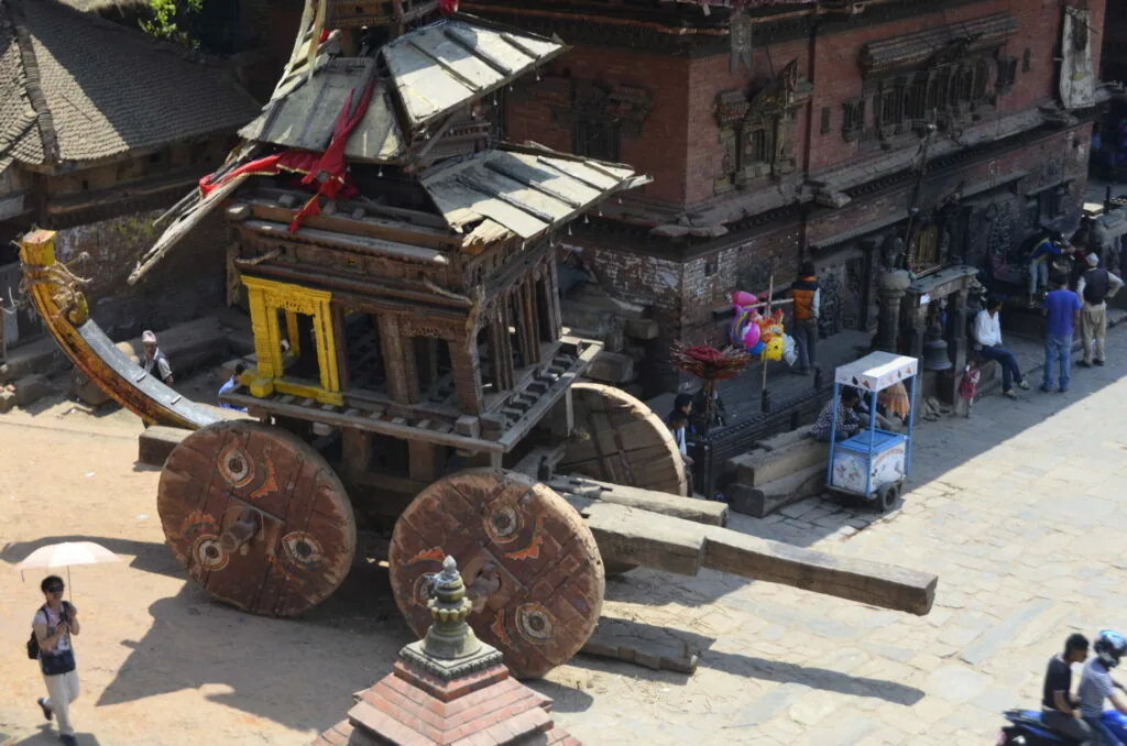 Ornate wooden chariot with large decorated wheels in historic courtyard with brick buildings and tourists