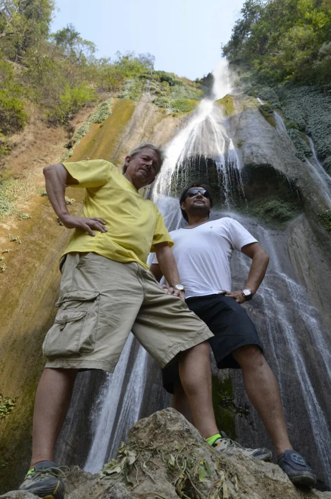 Two men posing in front of a cascading waterfall flowing down a rocky hillside covered with vegetation