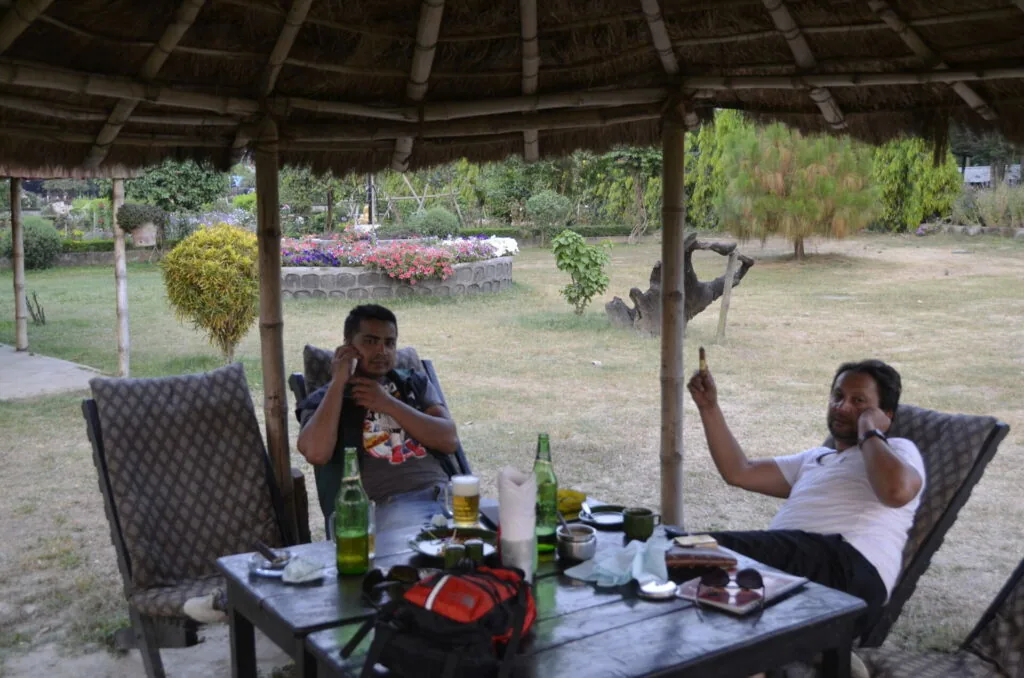 Two men relaxing under a thatched roof pavilion with beer bottles and food on a table, garden landscape visible in background