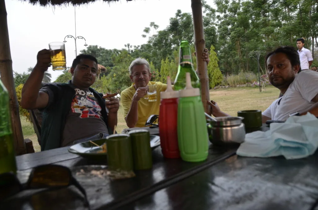 Group of men gathered around outdoor table with beverages and food under shelter with trees in background