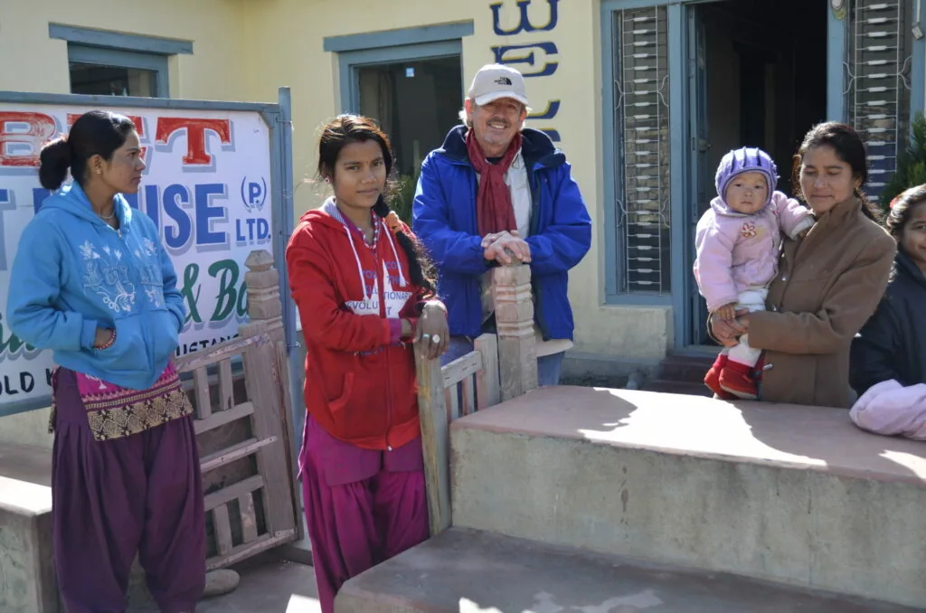 Group of people standing in a courtyard, including a man in a blue jacket and white cap interacting with locals, with a child being held on the right side.