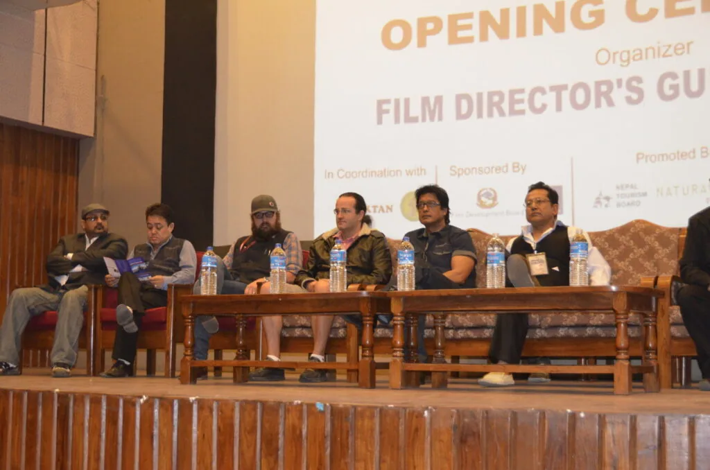 Panel of six men seated at a table during a film director's guide event, with water bottles in front of them and a banner visible behind.