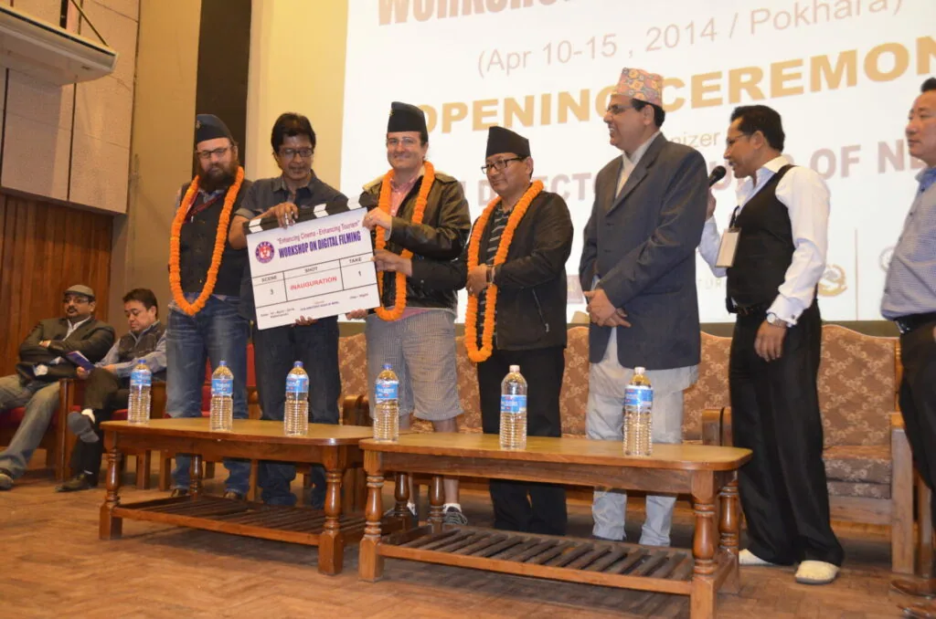 Officials and dignitaries stand on stage holding a ceremonial check during an opening ceremony event, wearing traditional garlands and formal attire.