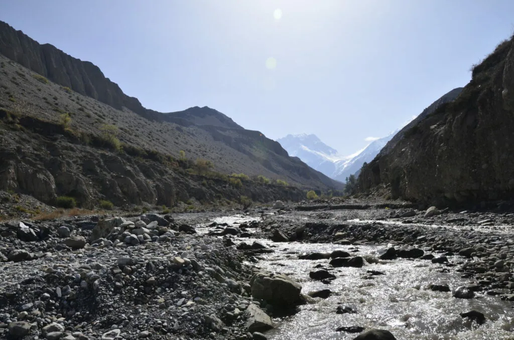 Mountain valley with steep rocky slopes, flowing river with boulders, snow-capped peak visible in distance under clear blue sky