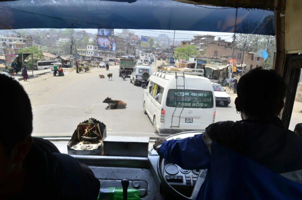 View from inside a bus showing a busy street scene in a Nepali town with buildings, vehicles, and pedestrians