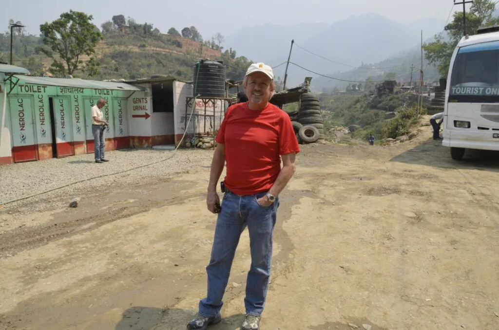 Man in red shirt and white hat standing on dirt road with public toilets and tourist van in mountainous area