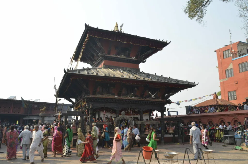 Crowds of people in colorful clothing gathered in a plaza in front of a multi-tiered traditional temple with ornate roofs and golden spires.