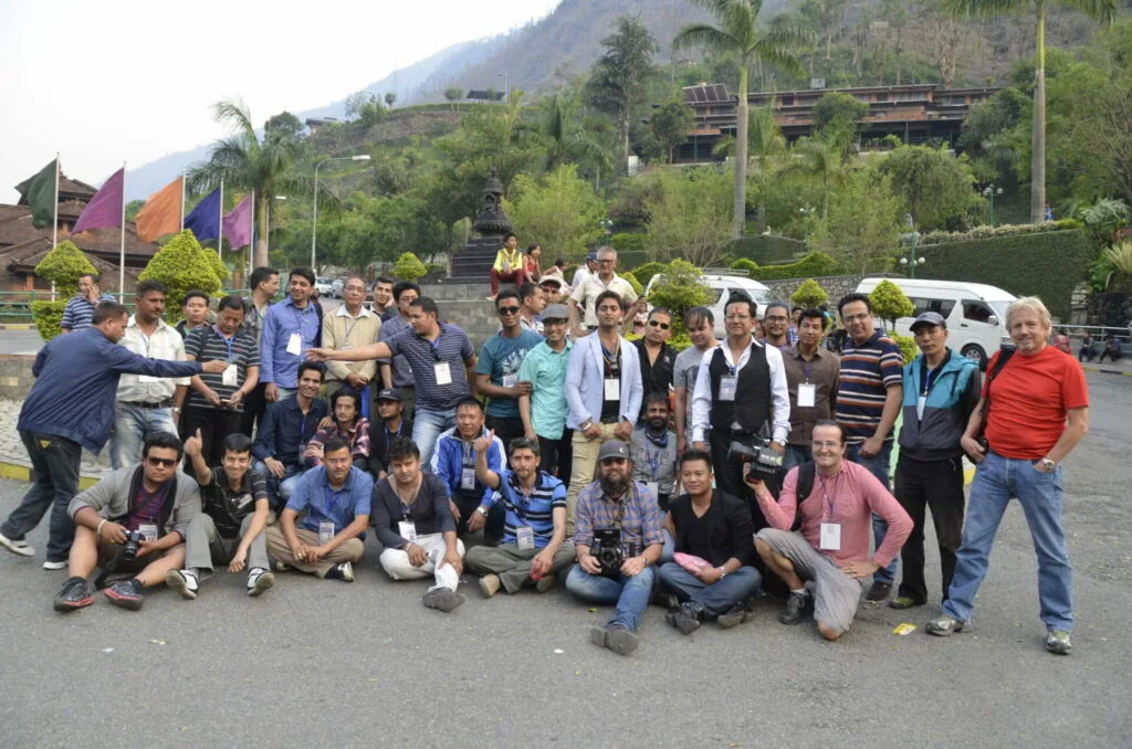 Large group of people posing together outdoors in front of a hillside with colorful flags and traditional architecture visible in background