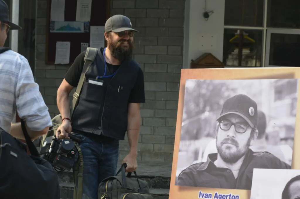 Bearded man wearing cap and dark vest holding camera equipment stands next to framed black and white portrait with name placard.