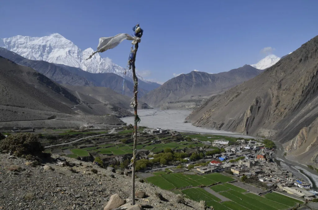 Prayer flag on wooden pole overlooking mountain valley village with snow-capped peaks and river