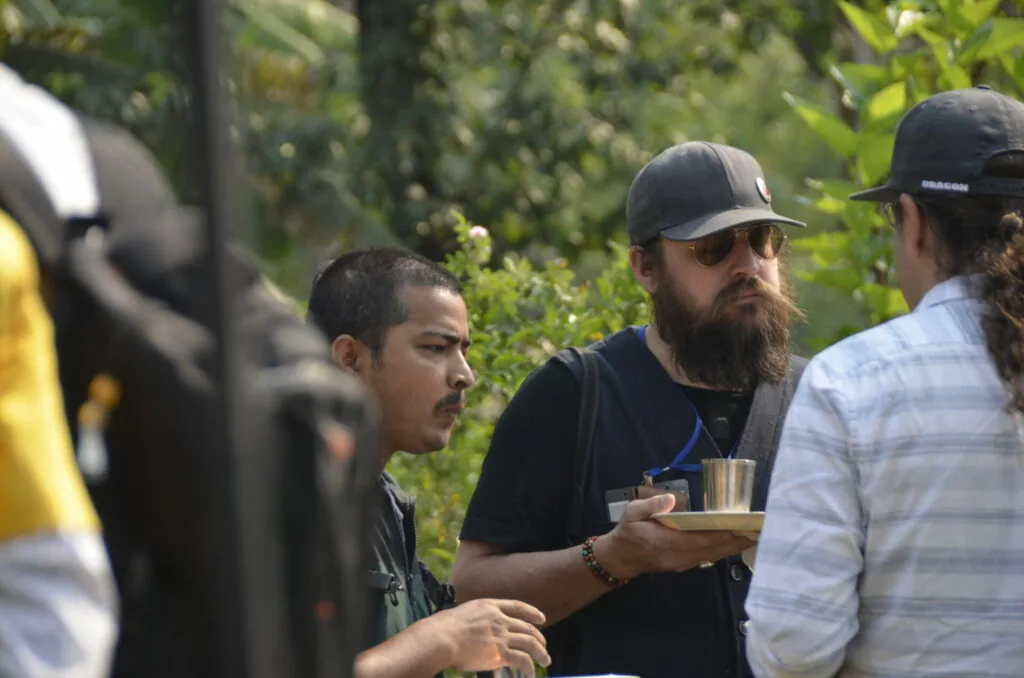 Three people conversing outdoors with green foliage in background, man in center wearing black shirt and cap holding a metal cup
