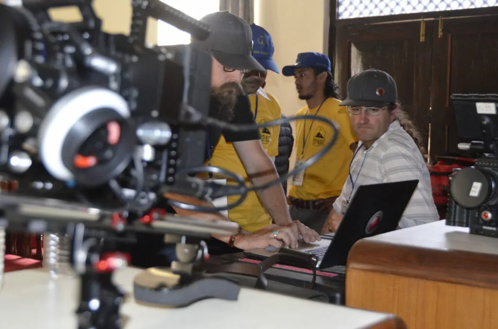 Film crew operating camera equipment indoors with three men in yellow shirts and caps, one viewing a laptop monitor