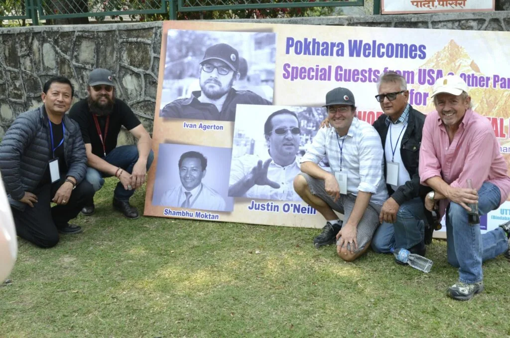 Six people posing with a welcome banner featuring photos of special guests from USA at what appears to be an event in Pokhara