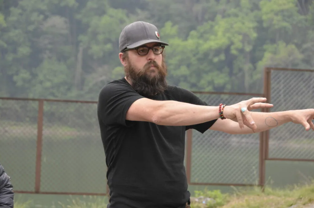 Bearded man in black shirt and gray cap stretching his arm at outdoor facility with trees and metal fence in background