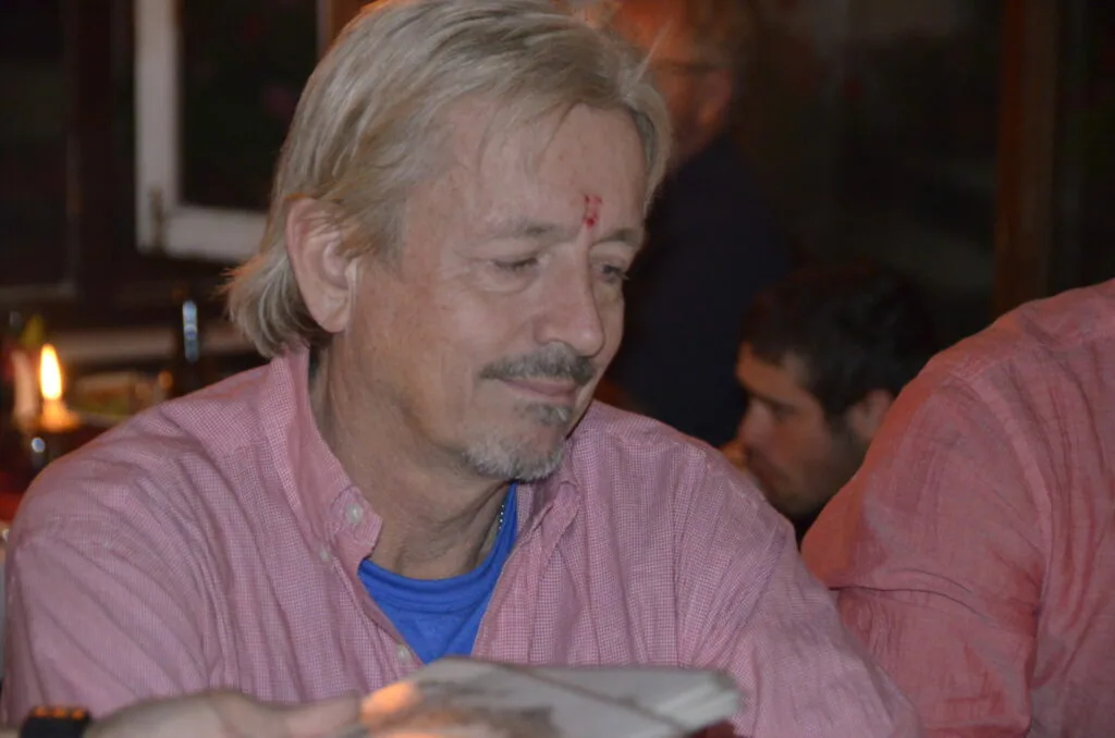 Middle-aged man with gray hair and mustache wearing a pink button-up shirt and blue undershirt at an indoor gathering