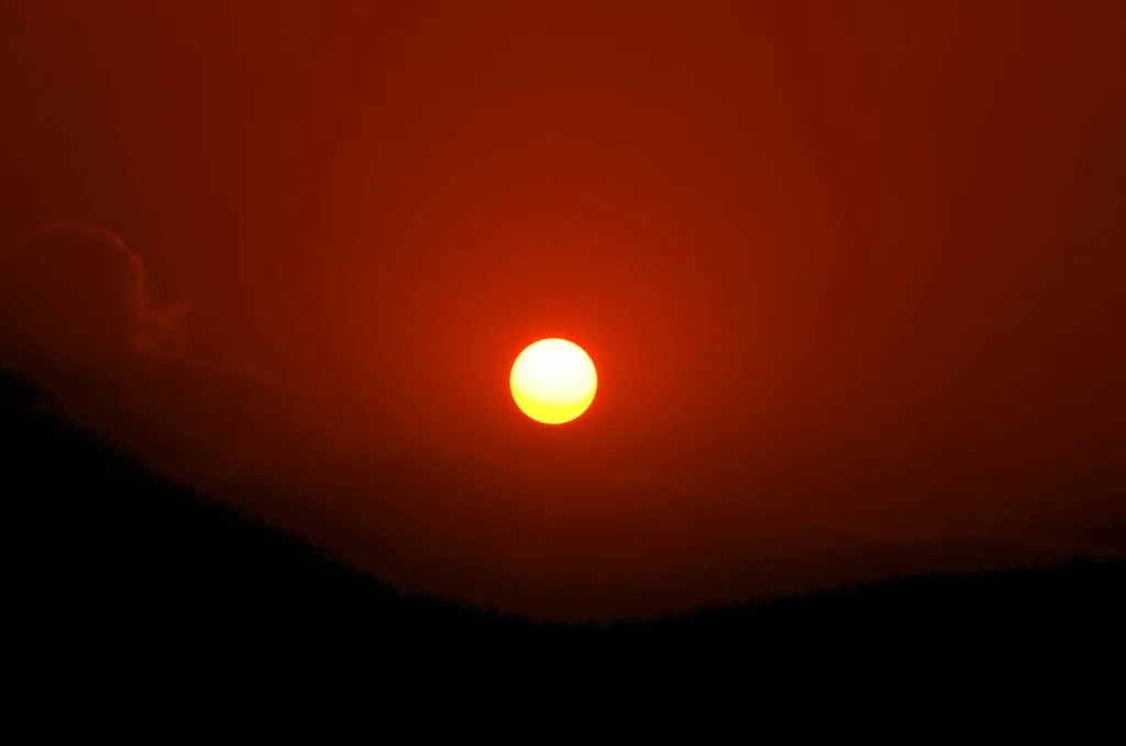 Bright sun setting on horizon above dark silhouetted landscape against orange and red sky