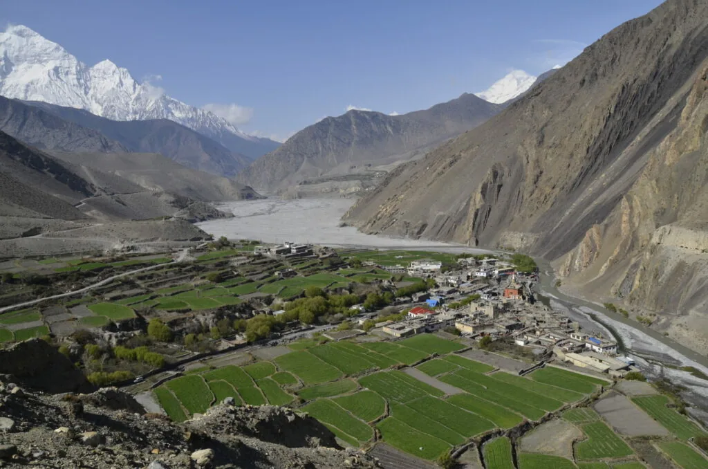 Aerial view of a mountain valley with terraced green fields, a river, village settlement, and snow-capped peaks under blue sky