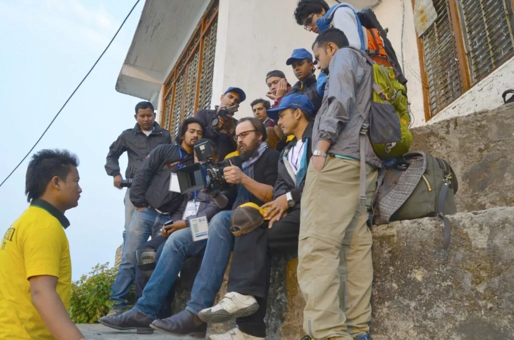 Group of men gathered outdoors, some wearing blue caps and lanyards, appearing to be at a training or community event near a concrete building.