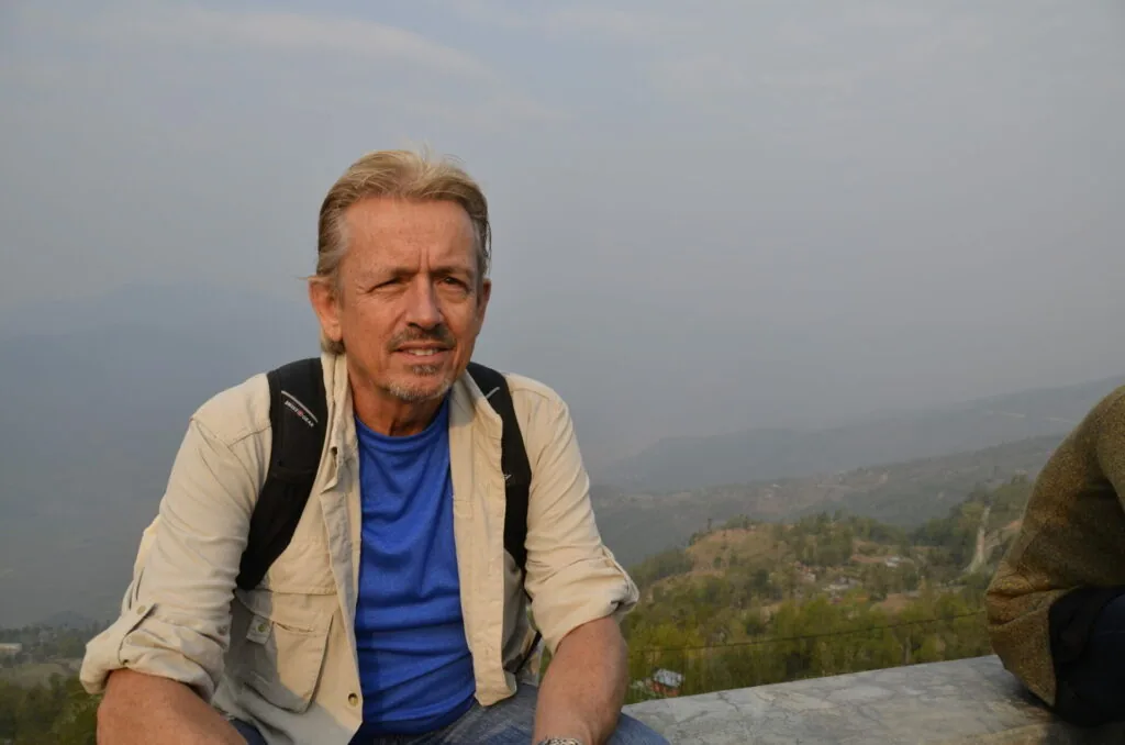Middle-aged man with light brown hair wearing a blue shirt and tan jacket with backpack straps, sitting outdoors with mountainous landscape in background