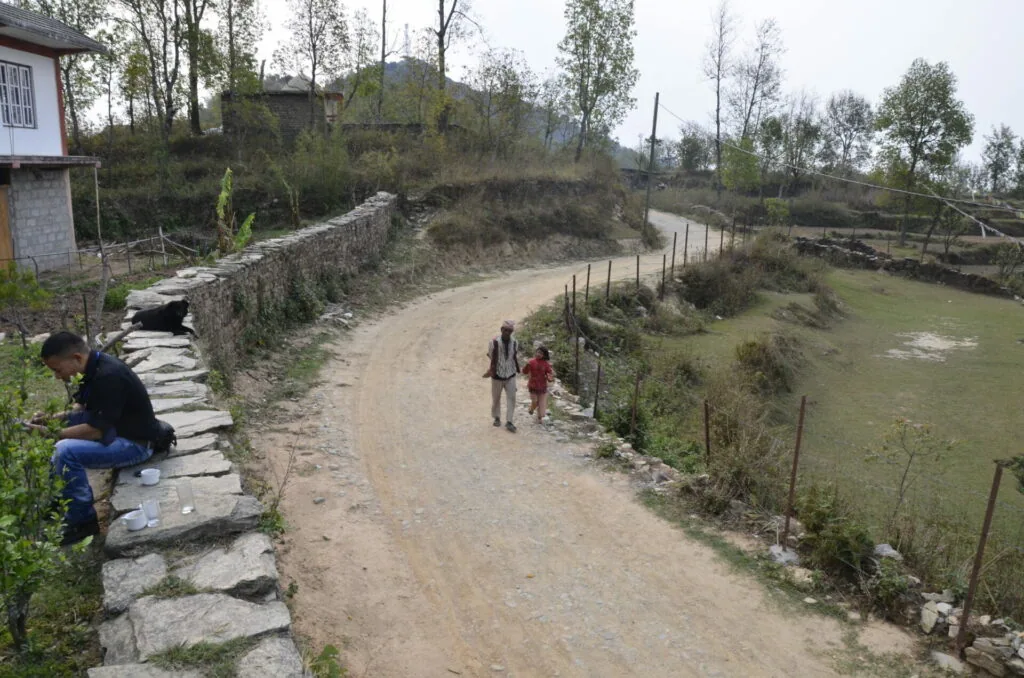 Rural village path with stone walls, two people walking on dirt road, agricultural fields and scattered trees in background