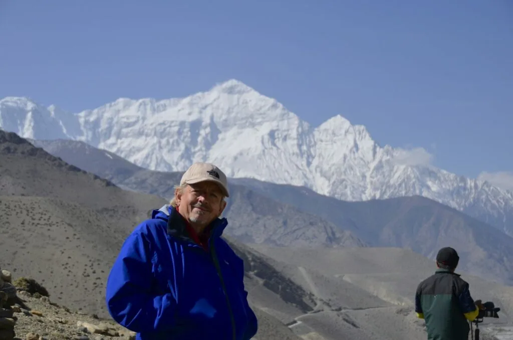 Man in blue jacket and cap standing on mountain slope with snow-covered peaks in background