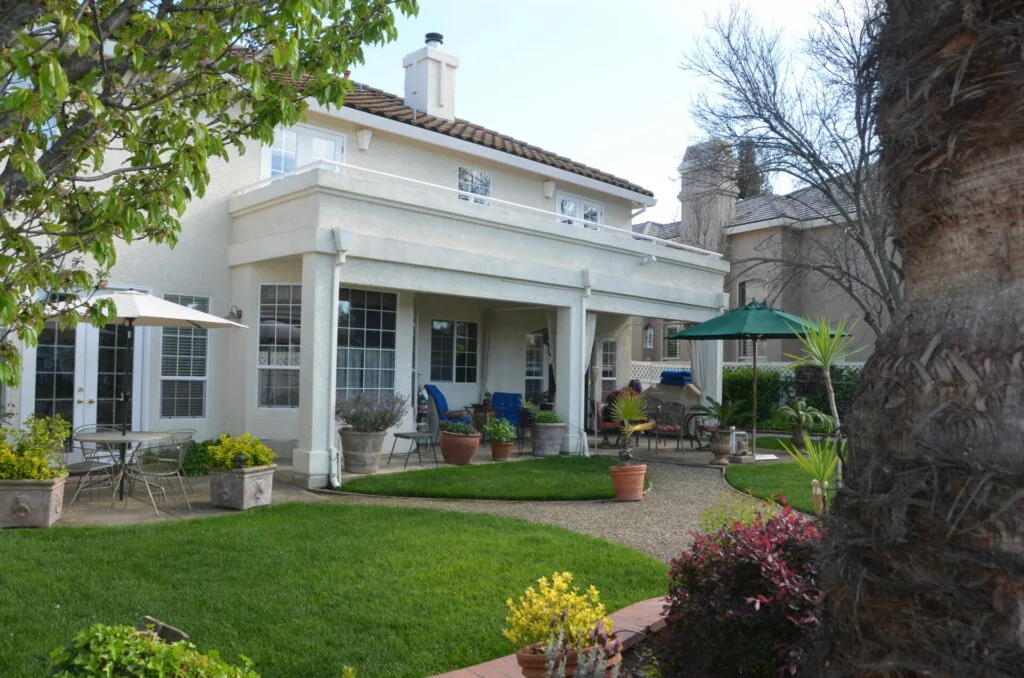 White two-story house with covered patio, green lawn, potted plants, and patio furniture with umbrella