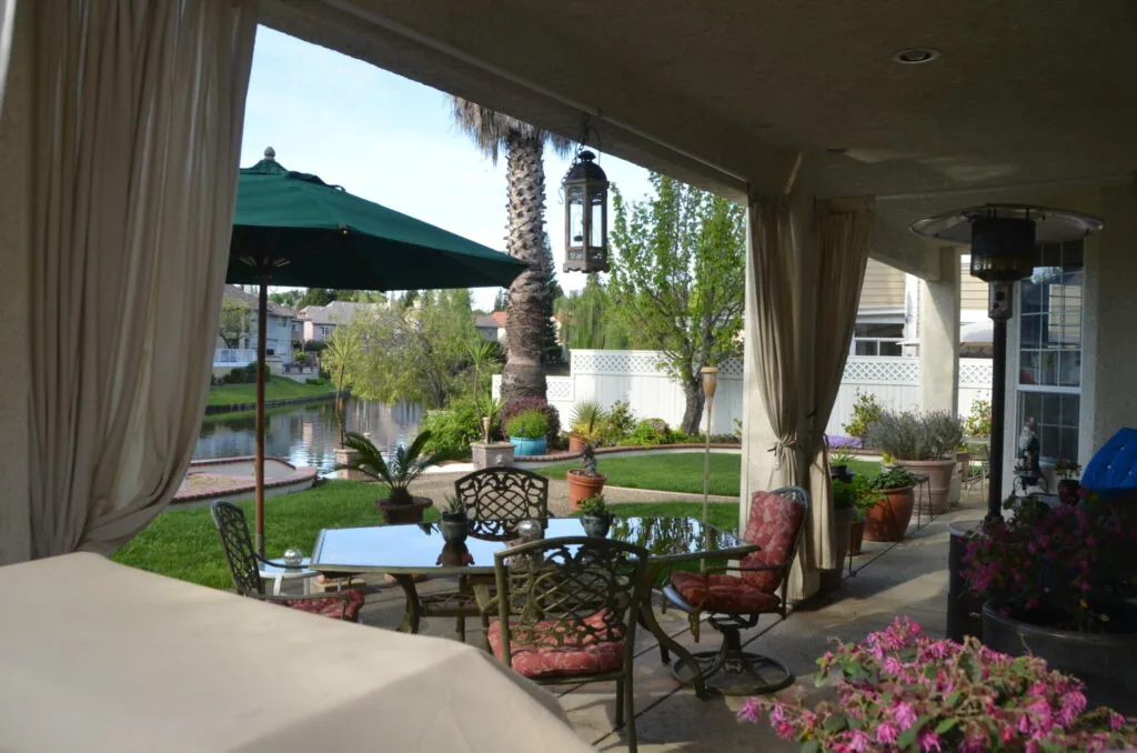 Covered patio with metal furniture, pink flowers, palm tree, and green umbrella overlooking a residential backyard with water feature