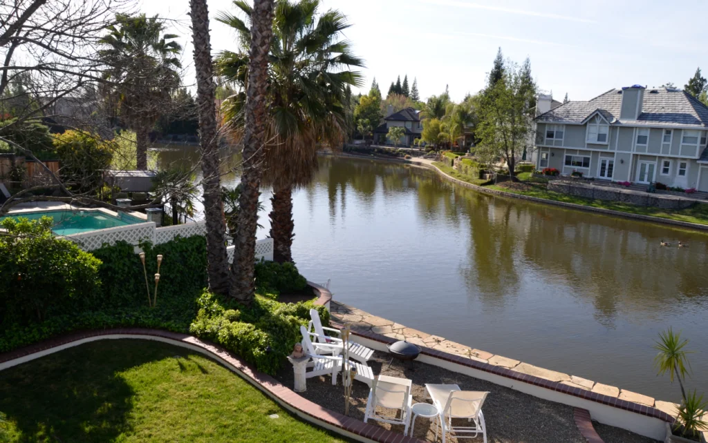 Waterfront property with palm trees, manicured lawn, seating area, and multi-story house across calm river