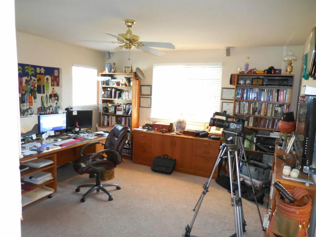 Home office and media room with wooden desks, computer setup, bookshelves, camera on tripod, and organized storage