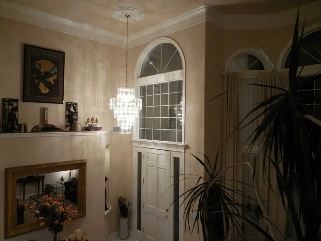 Elegant entryway with arched transom window, pendant chandelier, fireplace with mantel, potted plants, and framed artwork on cream-colored walls