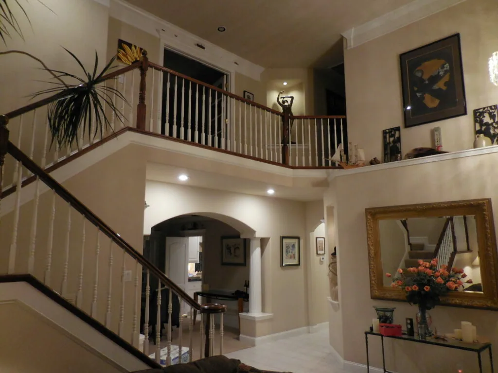 Two-story foyer with curved staircase, console table with mirror and flowers, artwork on walls, and potted plant on upper balcony