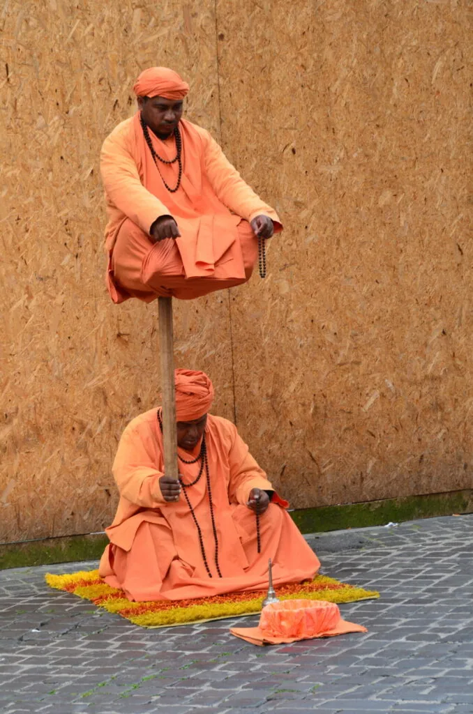 Two monks in orange robes performing acrobatic stunt against cork wall, one balanced on pole above the other