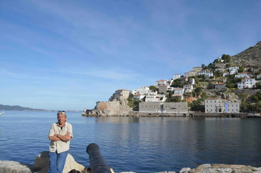 Man standing on rocky shore facing hillside village with white and stone buildings cascading down slope to blue Mediterranean water