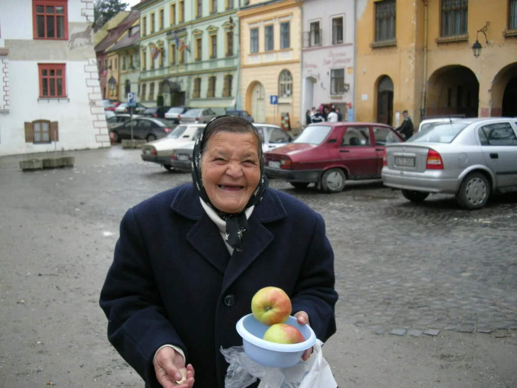 Elderly woman holding a bowl of apples in a European town square with colorful historic buildings and parked cars behind her
