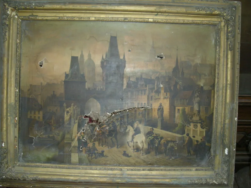 Framed painting depicting a medieval European city skyline with gothic towers and spires, a crowded town square below with figures and horses, in muted earth tones within a worn gilt frame.