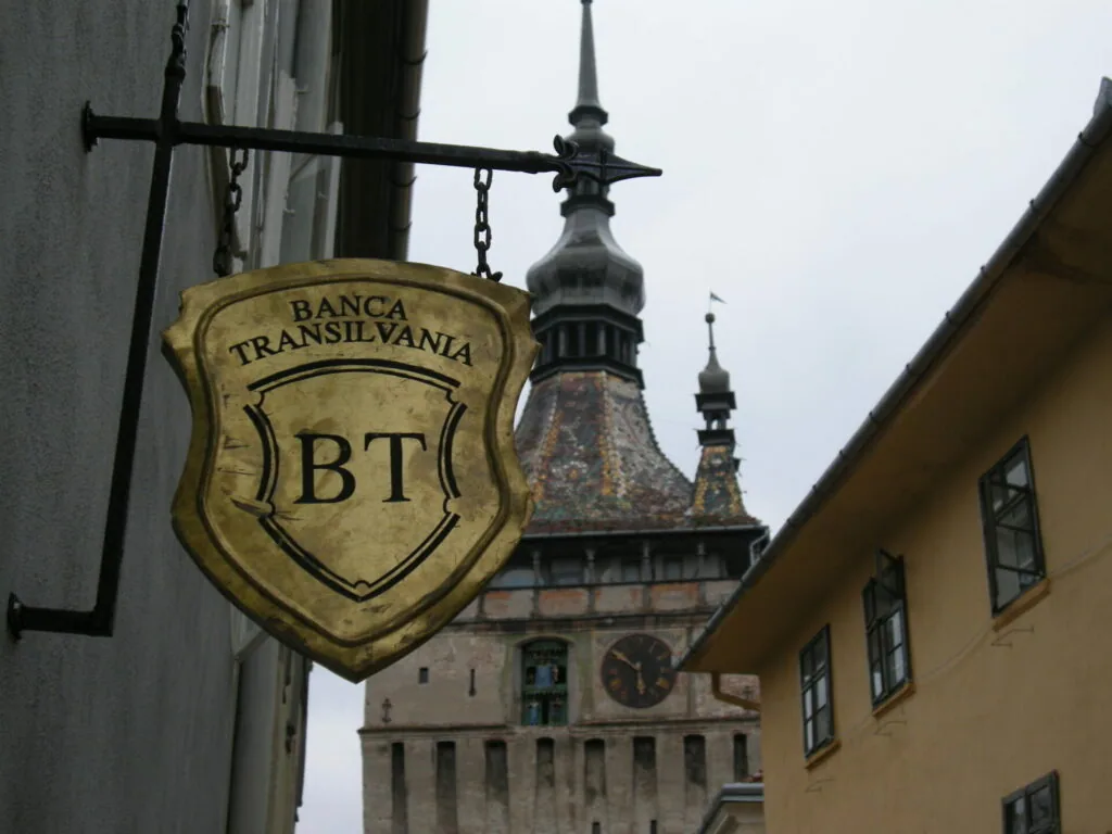Gold shield-shaped sign reading 'BANCA TRANSILVANIA BT' hanging from an iron bracket, with a medieval tower featuring a spire visible in the background
