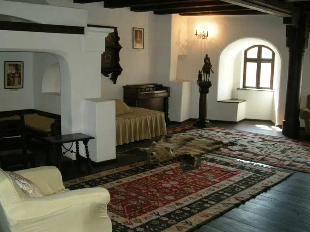 Interior of a room with white walls, dark wooden beams, ornate rugs, vintage furniture, and an arched doorway with a cross visible on the wall
