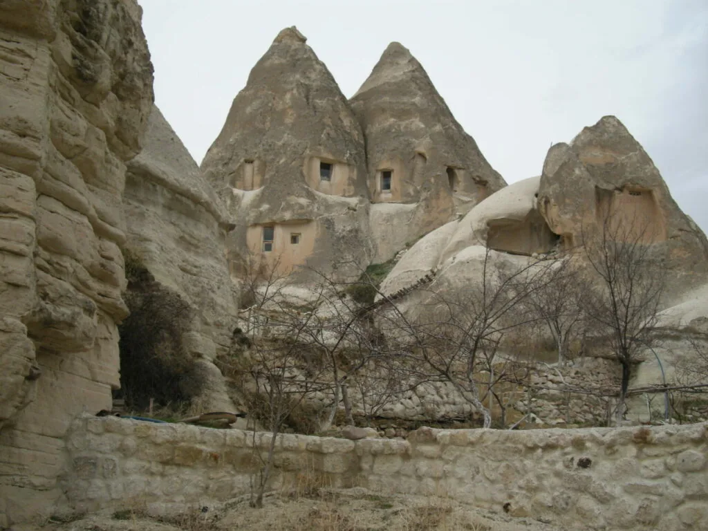 Cone-shaped rock formations carved with window openings and doorways, with bare trees growing at the base and stone wall in foreground