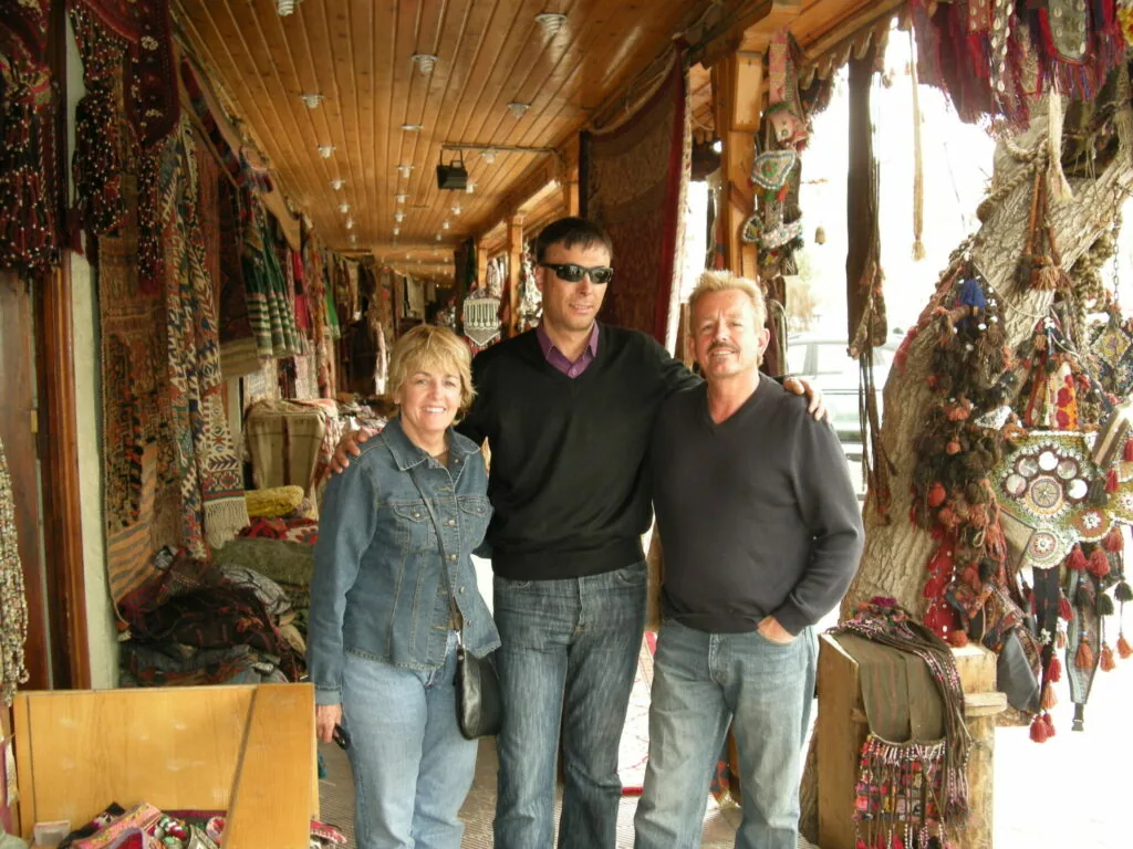 Three people standing in a covered market corridor lined with colorful traditional textiles and rugs hanging from both sides