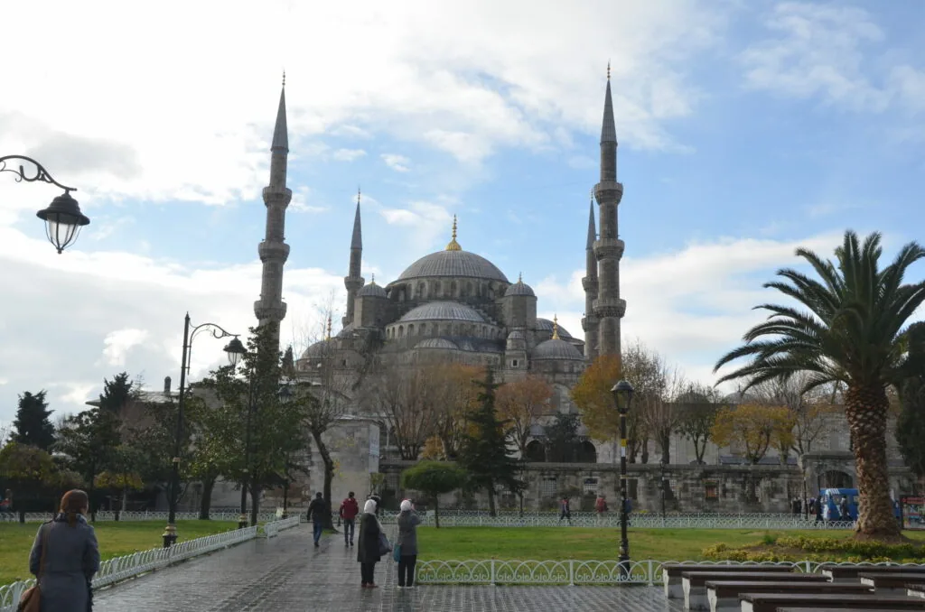Historic mosque with multiple minarets and central dome surrounded by park grounds with palm trees and visitors