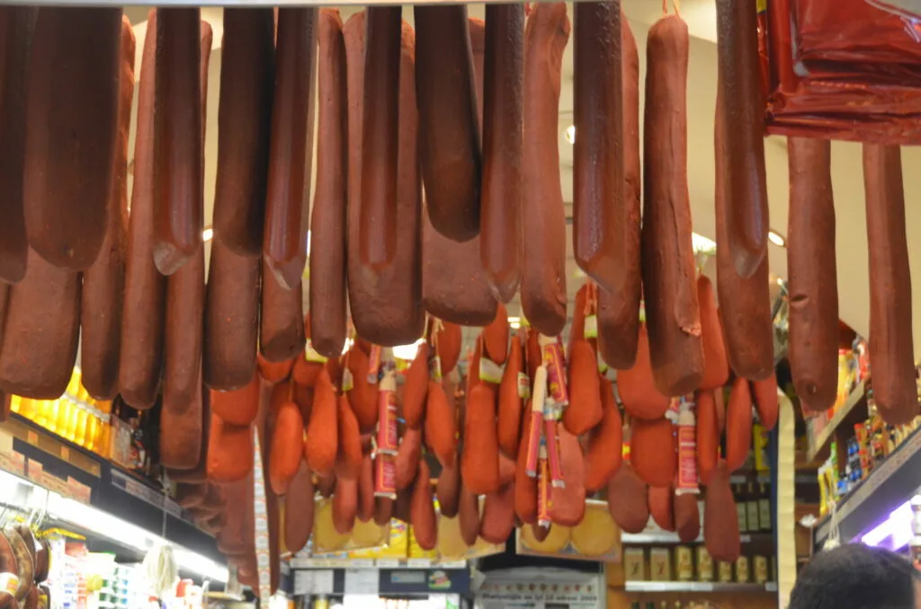 Cured meat products hanging from ceiling in market display, including sausages and other processed meats in various shades of red and brown.