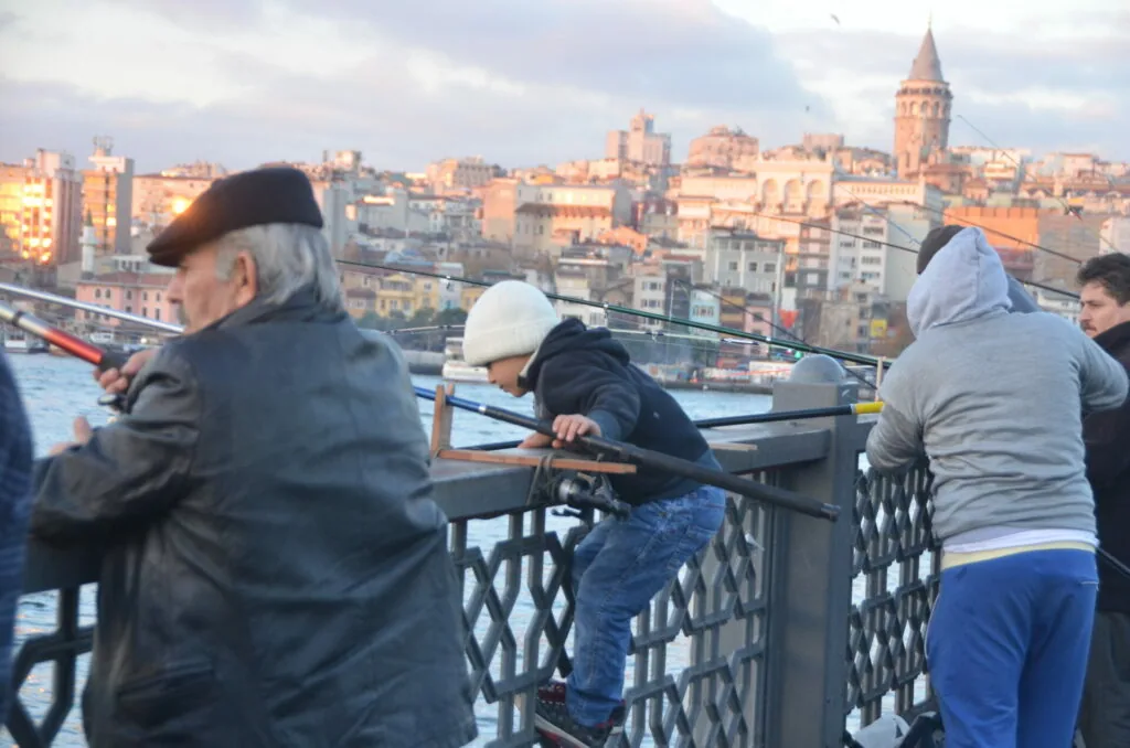 People fishing from a bridge railing overlooking a city skyline with a prominent tower in the background