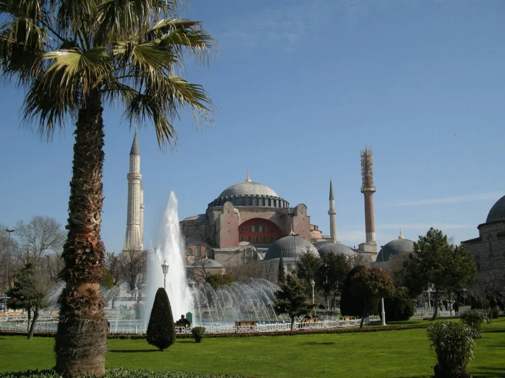 Historic mosque with large dome and minarets viewed across manicured green lawn with fountain and palm tree in foreground