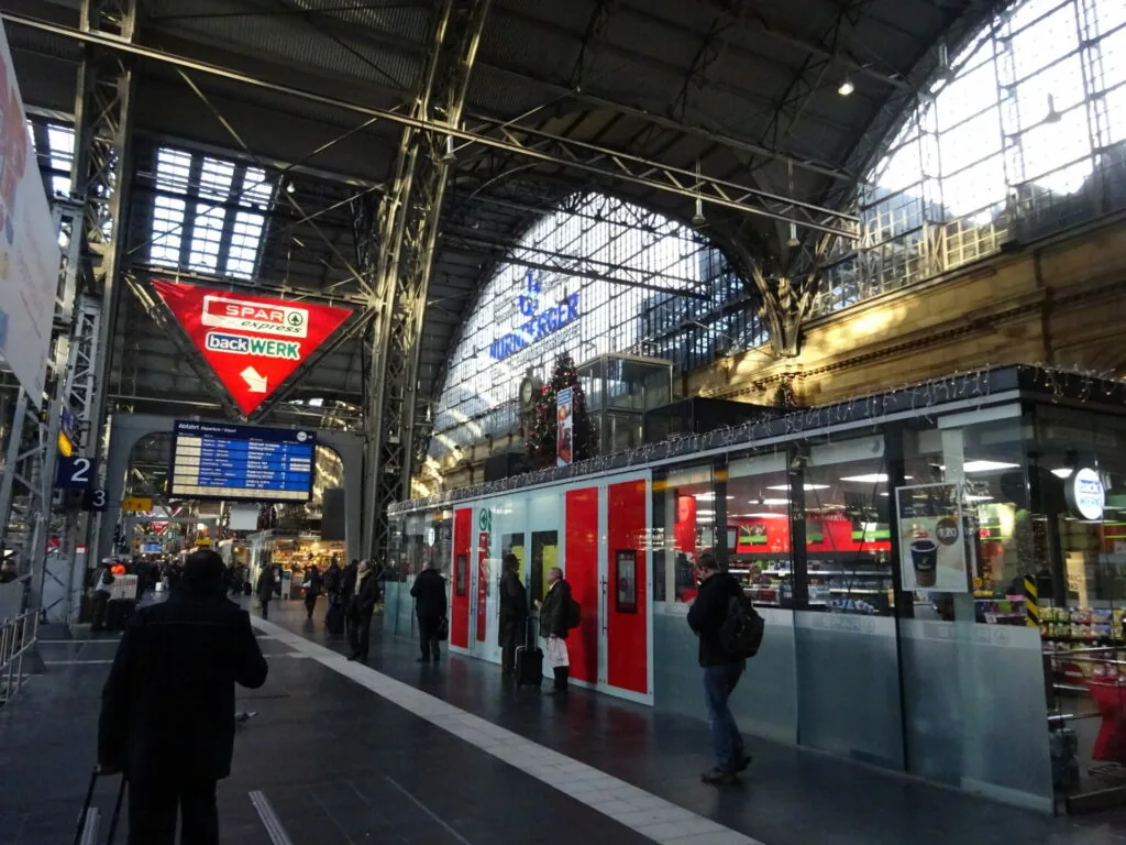 Train station platform with red and white train, industrial ceiling with metal beams and glass panels, retail storefronts and signage.