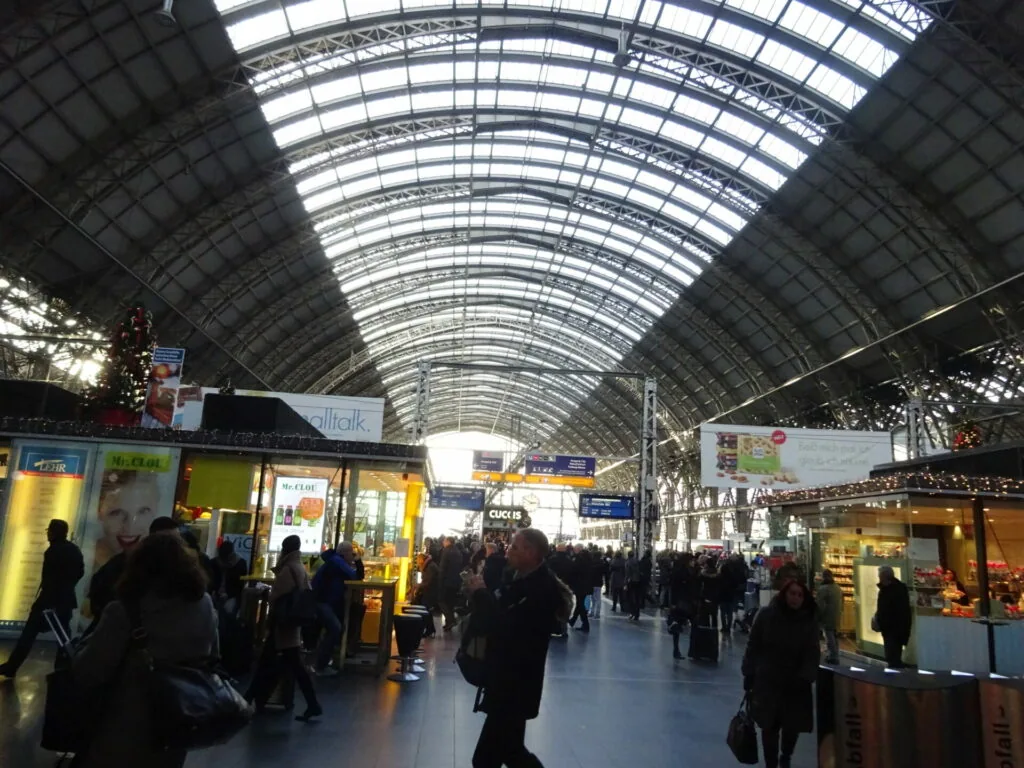 Interior of a busy train station with arched glass roof, illuminated shops, and crowds of travelers walking through the concourse.