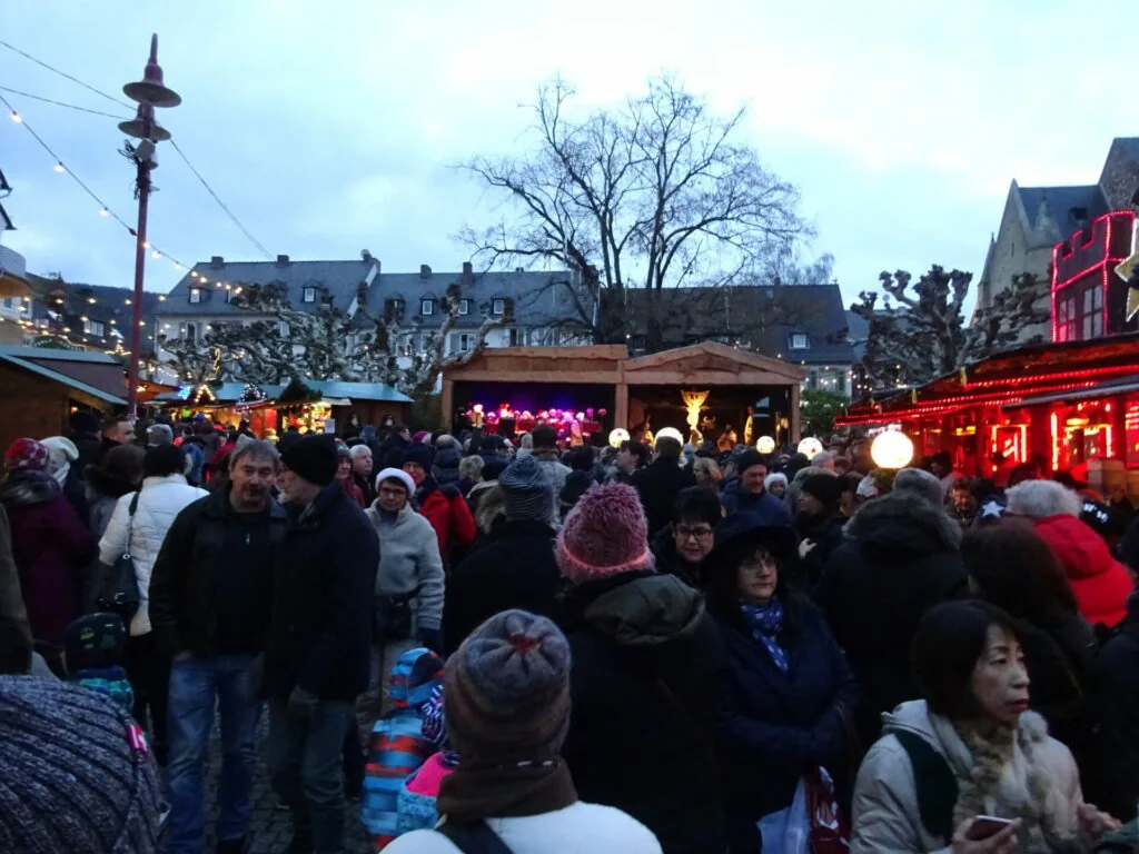 Crowded Christmas market at dusk with illuminated wooden stalls, historic buildings, and bare trees in background