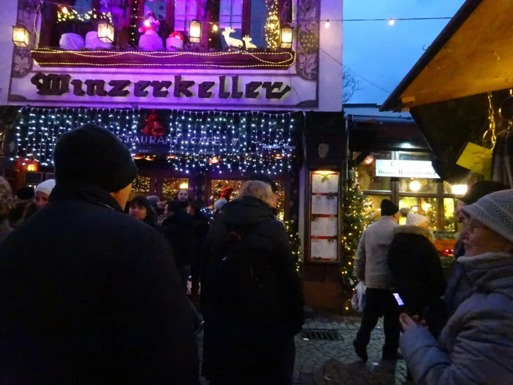 Festive market stall decorated with purple and white lights, crowds of people gathered at dusk during winter holiday market