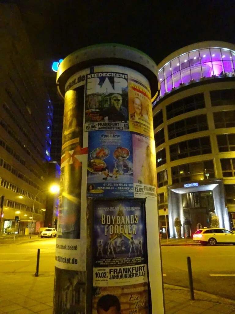 Cylindrical advertising column at night with multiple event and product posters, illuminated yellow lighting, modern buildings with purple-lit windows in background