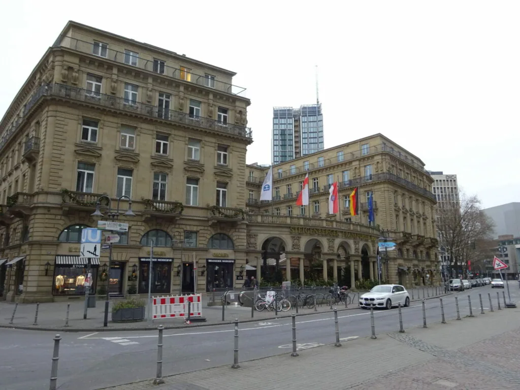 Historic European plaza with ornate classical buildings, flags hanging from facades, and modern high-rise in background
