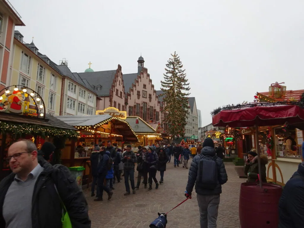 Crowded Christmas market in European city square with decorated wooden stalls, historic red building, and coniferous tree